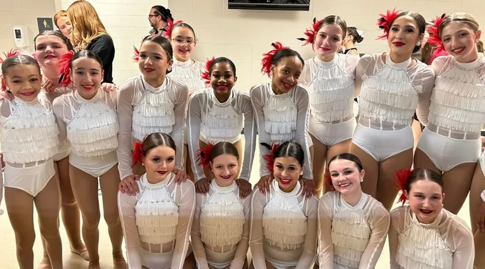 Un gruppo di giovani danzatrici posa insieme in costumi bianchi e beige nella sala danza della Kokomo Center Township Consolidated School Corporation.