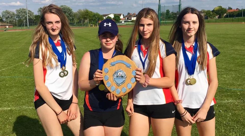 Quattro studentesse della Wisbech Grammar School tengono in mano medaglie e una coppa sul campo sportivo.