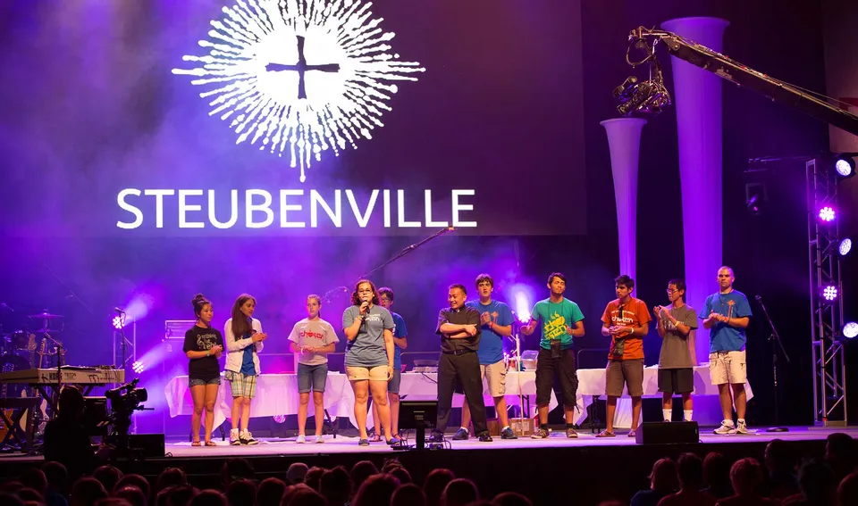 Studenti della Aquin High School sono in piedi sul palco davanti a una grande installazione luminosa a forma di croce con la scritta STEUBENVILLE.
