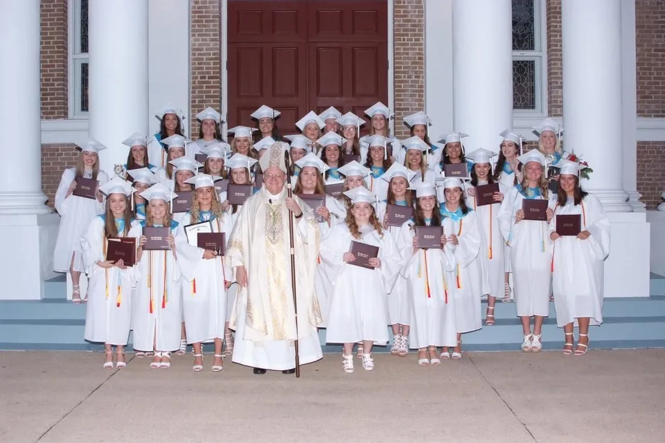 Studentesse della Our Lady Academy in toga bianca con diplomi posano con un religioso su una scalinata.