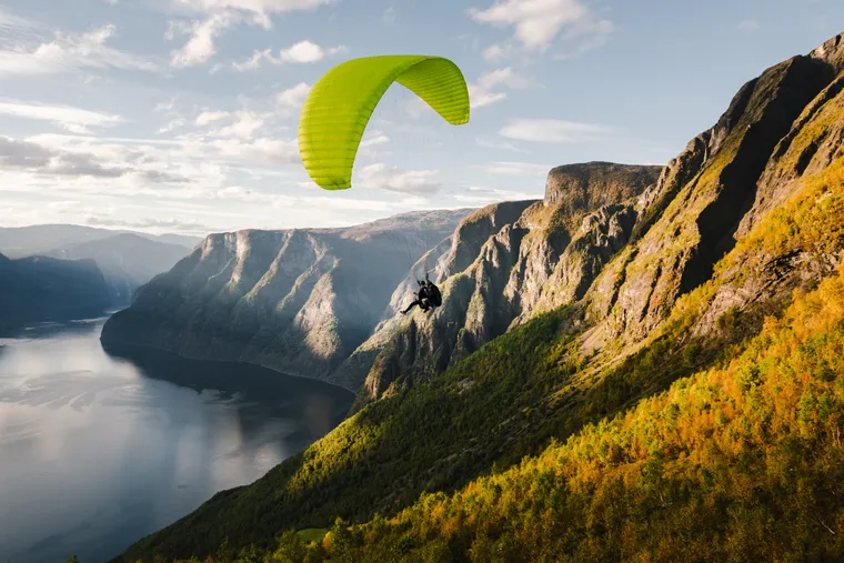 Gleitschirmflieger mit grünem Schirm über einem Fjord zwischen steilen Bergen