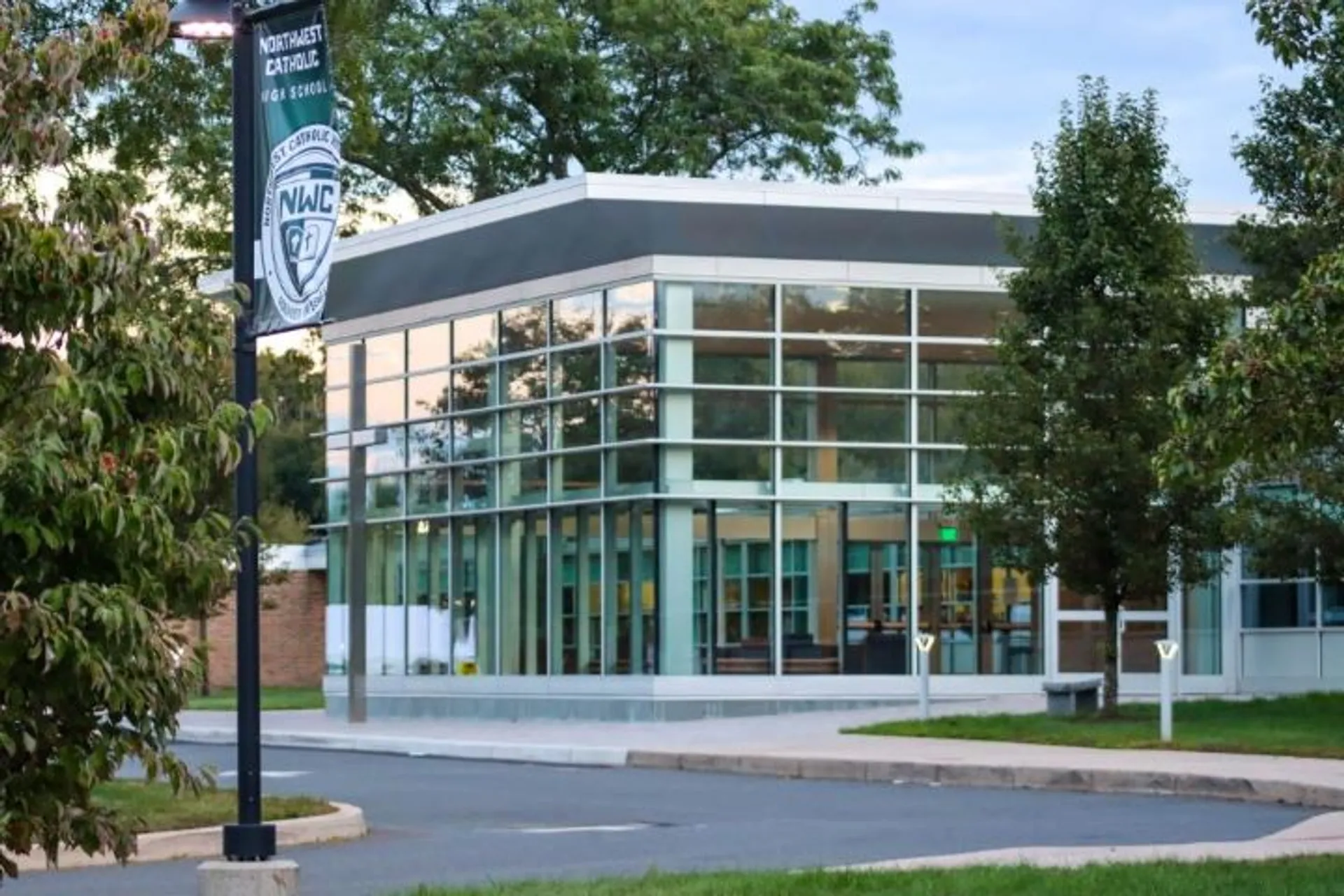 L'edificio moderno della Northwest Catholic High School, con facciata in vetro e logo, è circondato da alberi e da un vialetto pavimentato.
