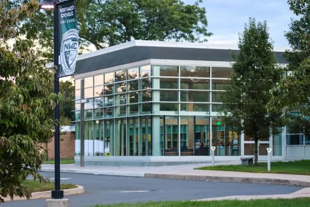 L'edificio moderno della Northwest Catholic High School, con facciata in vetro e logo, è circondato da alberi e da un vialetto pavimentato.