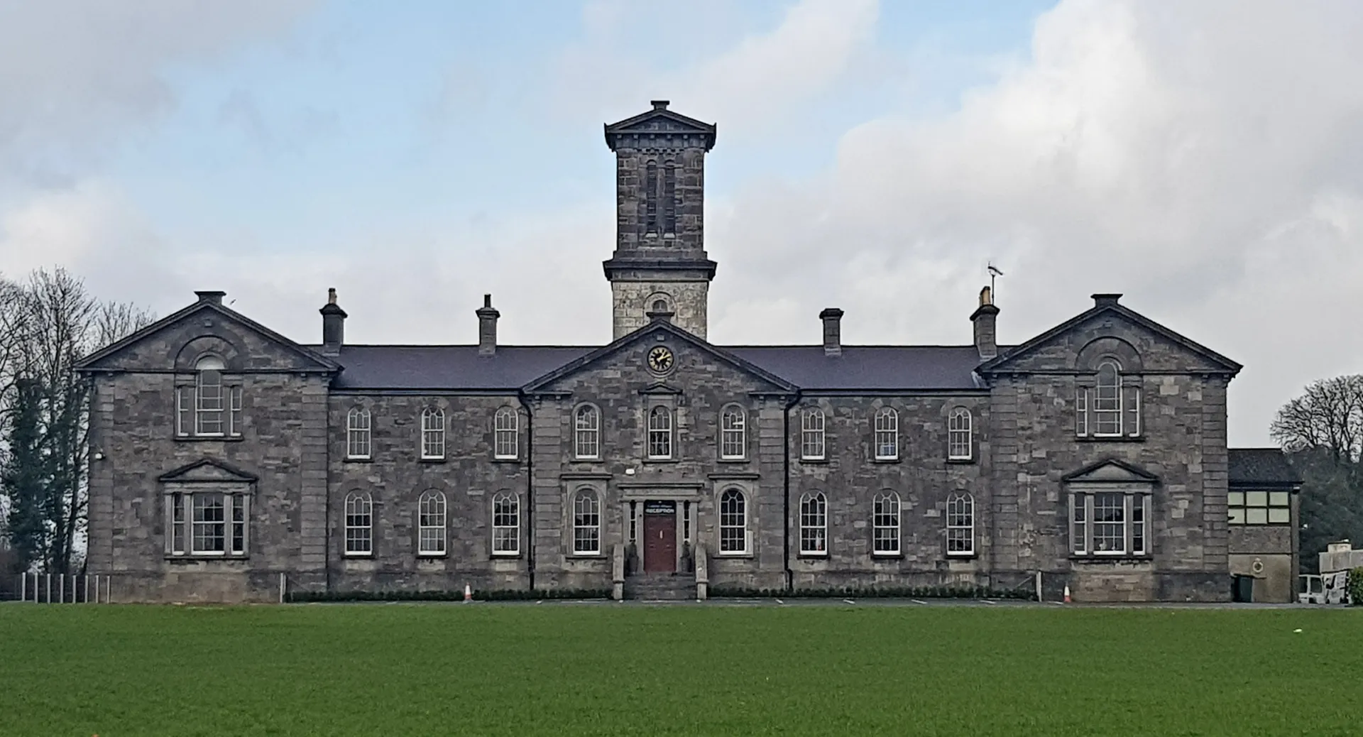 L'edificio con un'alta torre centrale del Coláiste Mhuire Mullingar sorge su un prato erboso sotto un cielo nuvoloso.