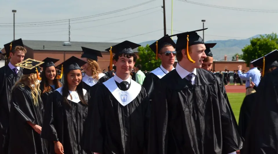 Un gruppo di diplomati della Bishop Kelly High School in toga nera si trova su un campo davanti a un edificio e alle montagne.