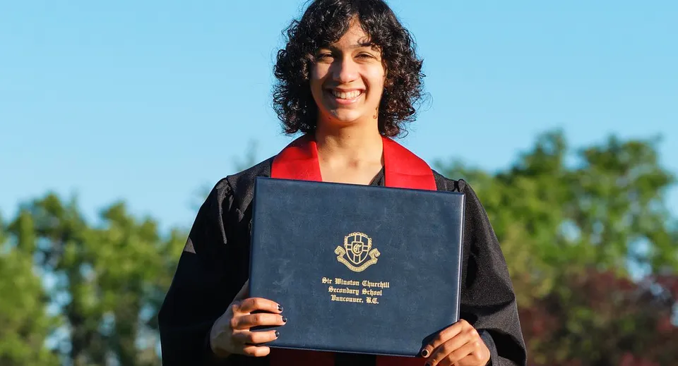 Uno studente della Sir Winston Churchill Secondary School indossa una toga da diploma nera e tiene in mano un diploma davanti a uno sfondo con alberi e cielo blu.