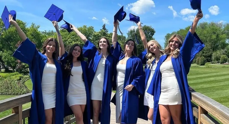 Un gruppo di diplomate del River East Collegiate festeggia all'aperto con le braccia alzate e i diplomi.