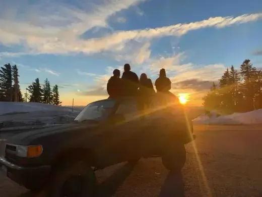 Quattro studenti seduti sul tetto di un pick-up guardano il tramonto in un paesaggio innevato