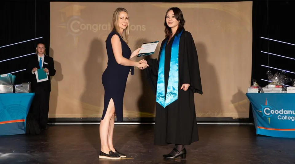 Due donne in abito formale si stringono la mano sul palco davanti a uno striscione di congratulazioni nel campus del Coodanup College.