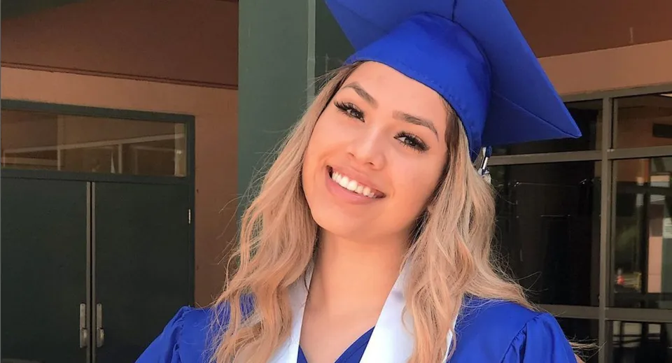 Una giovane donna con tocco e toga da diploma blu sorride davanti a una porta a vetri nel campus della Dr. Charles Best Secondary School.
