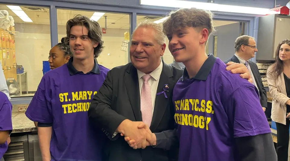 Tre persone in maglia viola accolgono un uomo anziano in abito elegante presso la St. Mary Catholic Secondary School.