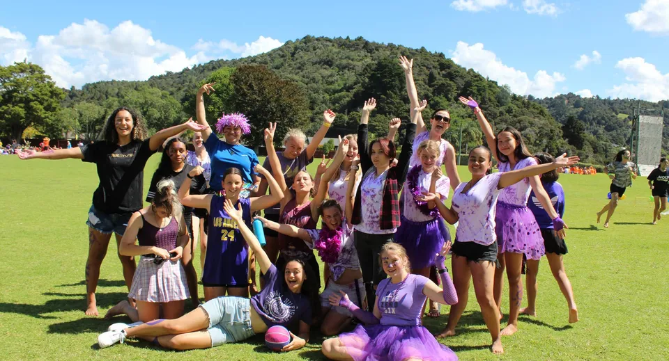 Un gruppo di studentesse della Whangarei Girls High School si trova su un campo erboso davanti a colline verdi sotto un cielo azzurro.