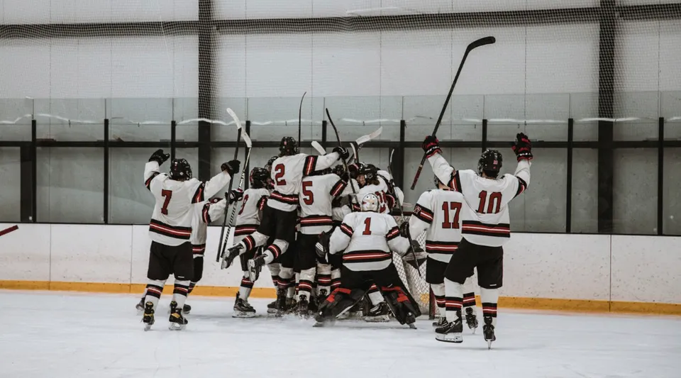 Un gruppo di giocatori di hockey della Pomfret School si raduna sul ghiaccio di una pista davanti a una parete di vetro.