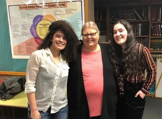 Una mujer adulta y dos jóvenes sonriendo juntas frente a un póster educativo y estanterías