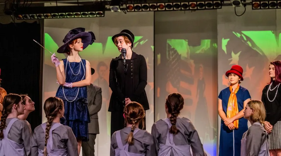Le studentesse della Girls Preparatory School mettono in scena uno spettacolo teatrale con costumi storici su un palco con illuminazione colorata.