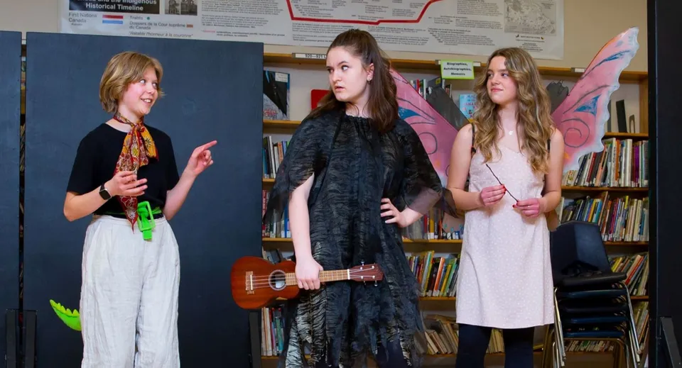 Tre studentesse del Daniel McIntyre Collegiate Institute con un ukulele in una stanza con scaffali di libri.