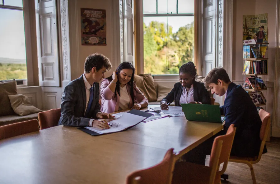 Cuatro alumnos y alumnas de Stover School trabajan juntos en tareas en una gran mesa de madera en una sala luminosa con ventanas de cuarterones.