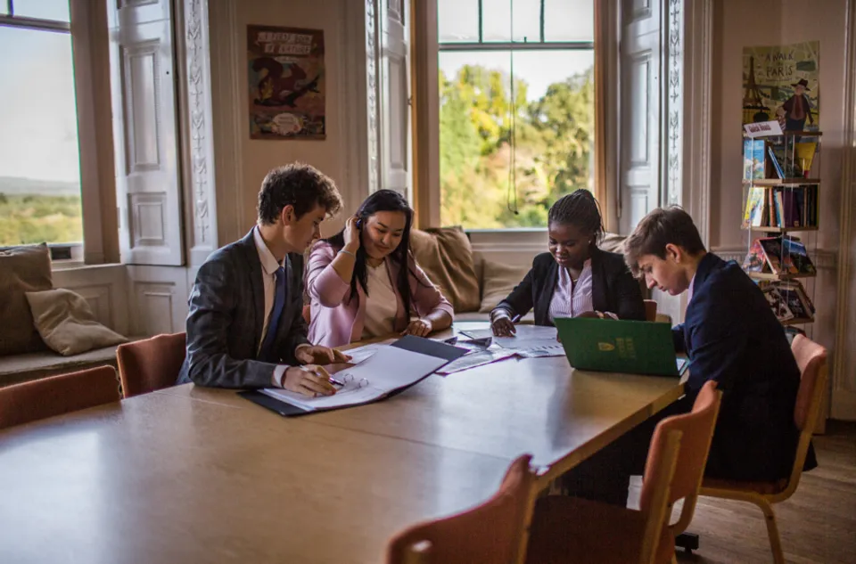 Quattro studentesse e studenti della Stover School lavorano insieme ai compiti a un grande tavolo di legno in una stanza luminosa con finestre a riquadri.