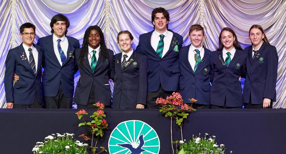 Studenti del Paraparaumu College in uniforme scolastica su un palco con una composizione floreale e un sipario sullo sfondo.