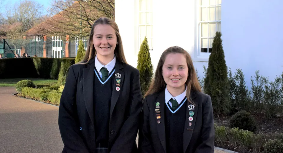 Due studentesse della Manor House School sono insieme all'aperto e sorridono davanti a un complesso edilizio con alberi.
