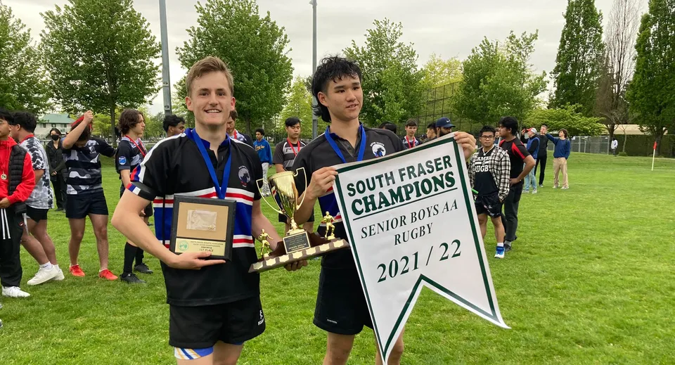 Due studenti della Clayton Heights Secondary School tengono una coppa e uno striscione di campionato all'aperto davanti a una folla.