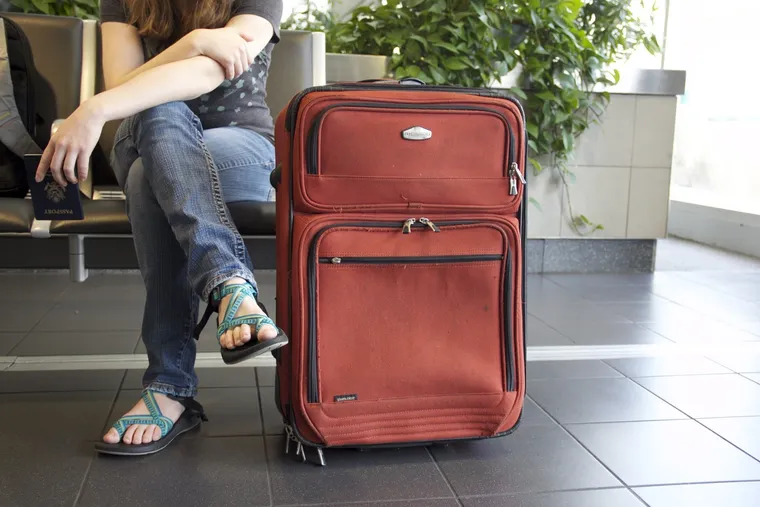 Person mit Reisepass in der Hand sitzt neben einem roten Rollkoffer im Wartebereich