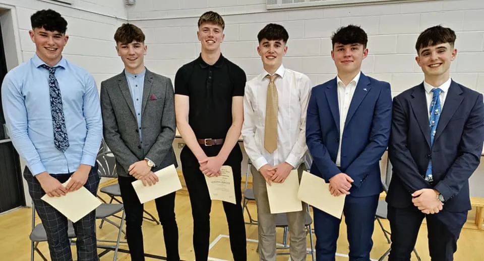 Un gruppo di sei giovani ragazzi in abito elegante si trova insieme davanti a uno sfondo bianco nel campus del St. Mary’s College.