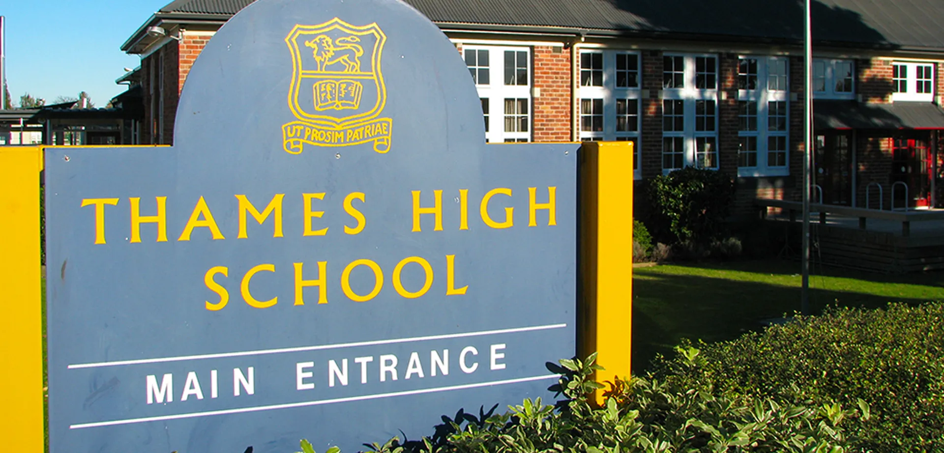 Il grande cartello della Thames High School con lo stemma davanti all'edificio in mattoni e all'area esterna curata.