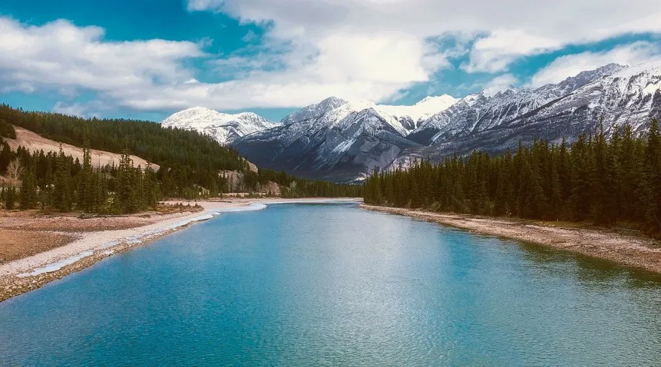 Un calmo fiume turchese scorre attraverso un paesaggio montano con cime innevate sotto un cielo nuvoloso.