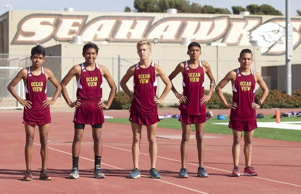 Cinque giovani atleti in maglia rossa si trovano sulla pista d'atletica della Ocean View High School davanti a un grande cartello SEAHAWK.