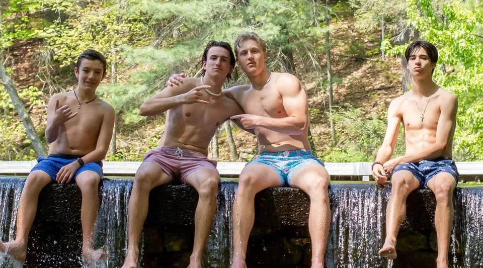 Quattro giovani uomini seduti a torso nudo su un muro con vista su un paesaggio boschivo nel campus della Avon Old Farms School.