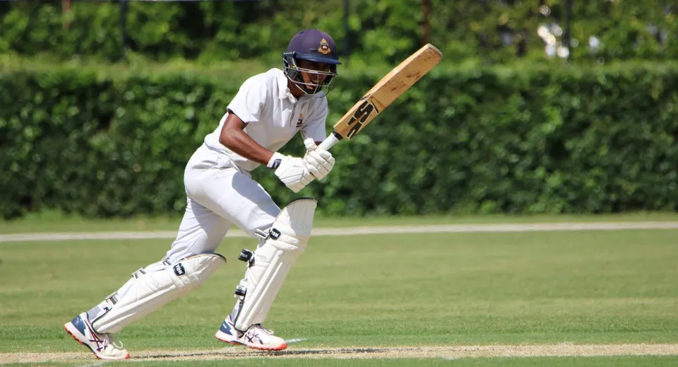 Un giocatore di cricket in divisa bianca tiene una mazza sul campo da gioco del St. Columba's College.