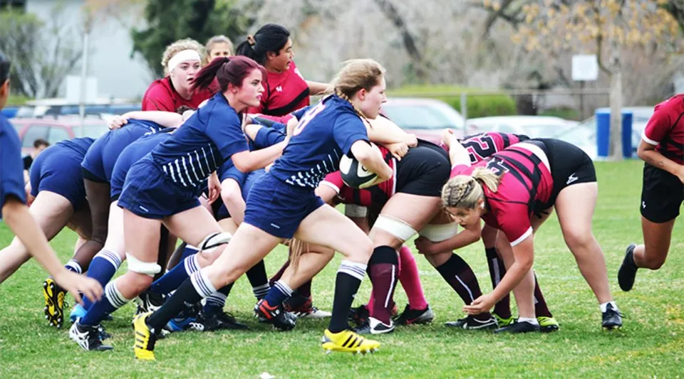 Studentesse della Crescent Heights High School durante una mischia di rugby su un campo d'erba con alberi ed edifici sullo sfondo.