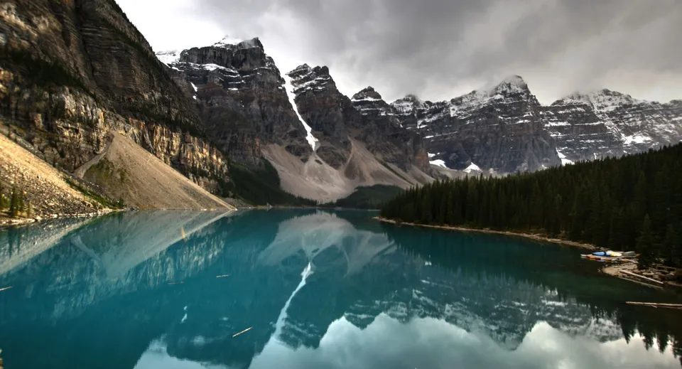 Un lago di montagna turchese riflette cime rocciose innevate e una foresta di conifere sotto un cielo nuvoloso.
