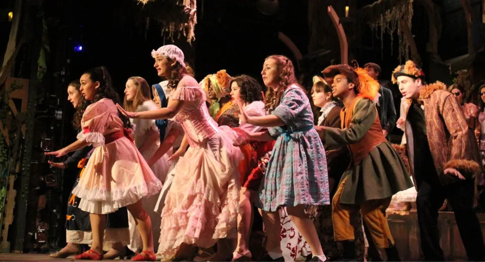 Un gruppo di studenti della Holy Cross High School presenta sul palco una vivace scena teatrale in costume.
