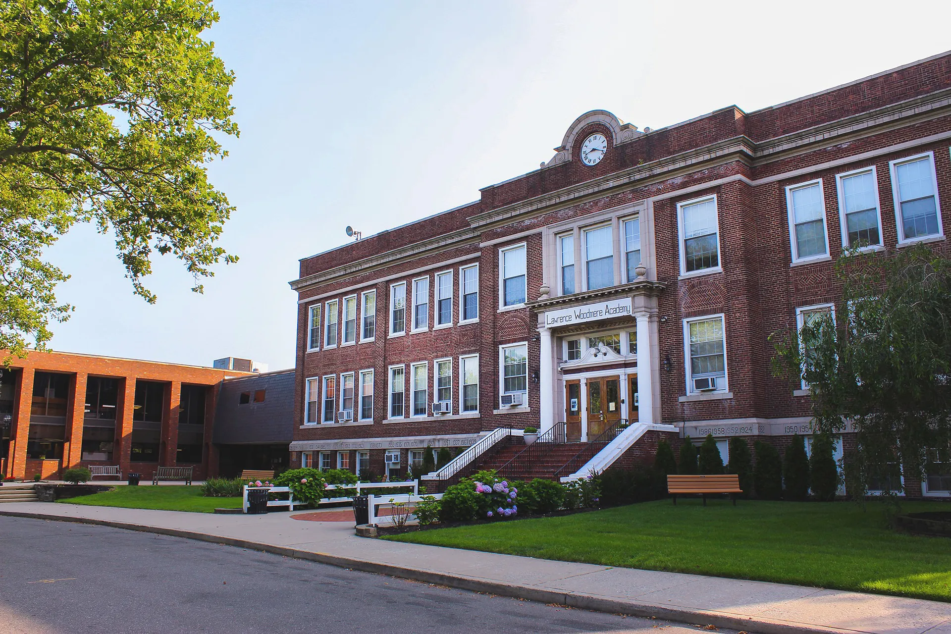L'edificio in mattoni a vista con torre dell'orologio nel campus della Lawrence Woodmere Academy è circondato da alberi verdi e da un vialetto pavimentato.