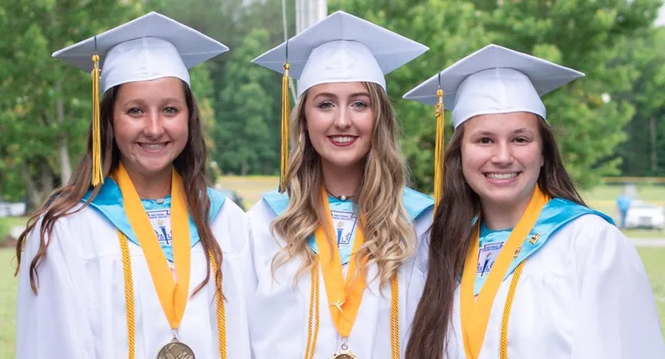 Tre studentesse della Kenston Forest School sono in piedi sorridenti nelle loro toghe da diploma davanti a uno sfondo verde.