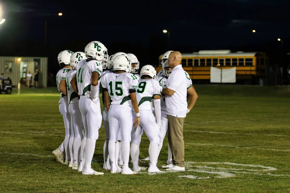 Studenti della Phoenix Christian Preparatory School in maglia bianca si trovano insieme su un campo da calcio scarsamente illuminato con uno scuolabus sullo sfondo.