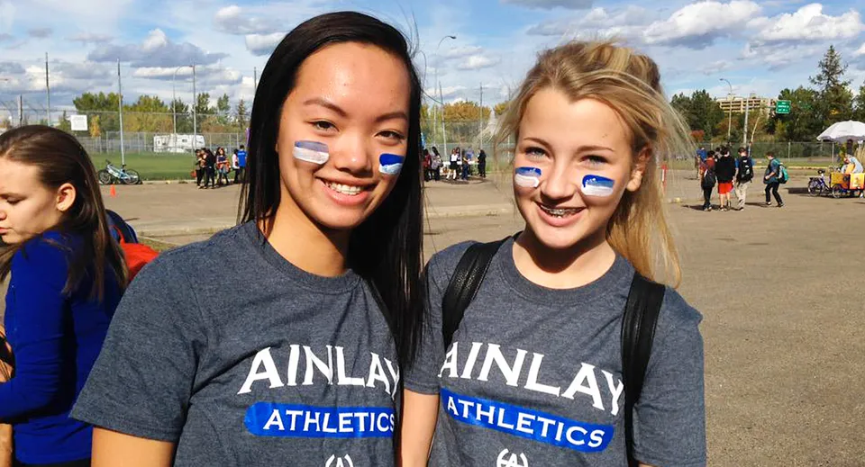 Due studentesse della Harry Ainlay High School sorridono davanti a un campo sportivo con altre persone sullo sfondo.