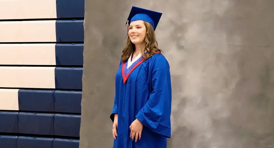 Una diplomanda in toga blu sta in piedi davanti a una parete grigia nel campus della Langley Secondary School.