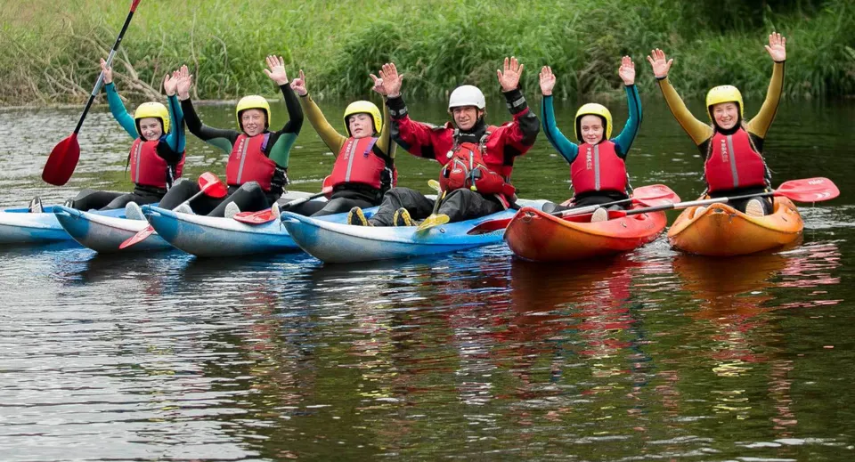 Un gruppo di studenti dell'Athlone Community College pagaia in kayak con giubbotti salvagente colorati su uno specchio d'acqua circondato da una verde vegetazione.
