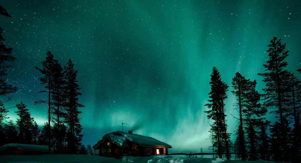 Un'aurora boreale verde brilla nel cielo stellato sopra una baita di legno innevata e alberi.