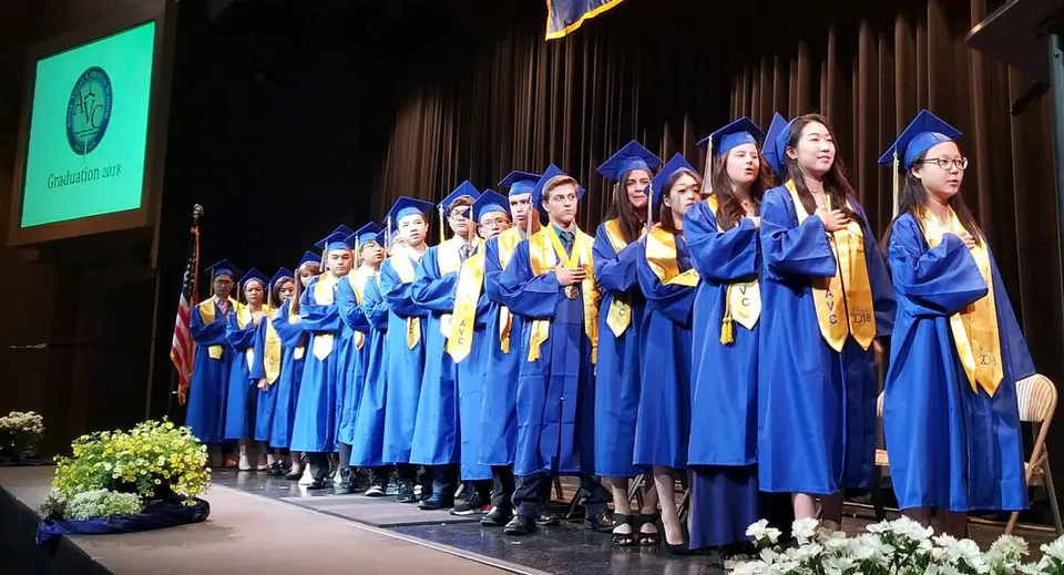 Un gruppo di diplomati della Apple Valley Christian School in toga blu su un palco davanti a un logo verde.