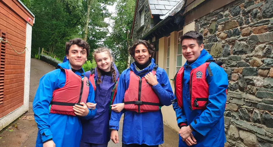 Quattro giovani della St. Bees School posano con giacche impermeabili blu e giubbotti di salvataggio rossi davanti a un edificio.