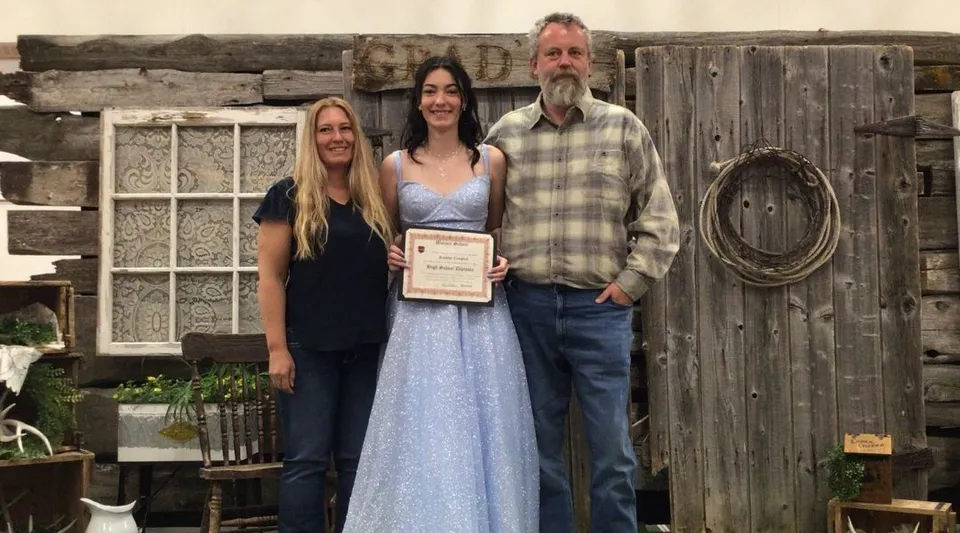 Una diplomata della Warner School posa con il suo diploma tra due adulti davanti a una parete di legno rustica.