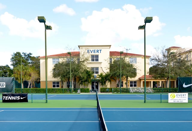 Ein blauer Tennisplatz im Vordergrund und das mehrstöckige Gebäude der Evert Tennis Academy im Hintergrund.
