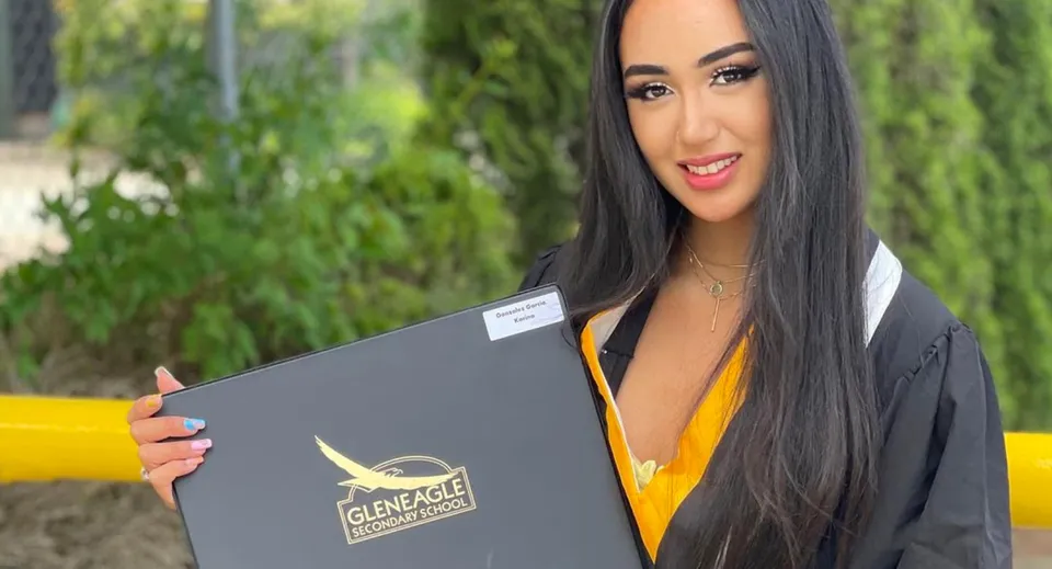 Una giovane donna tiene sorridente una cartella grigia nel campus della Gleneagle Secondary School davanti a uno sfondo verde.