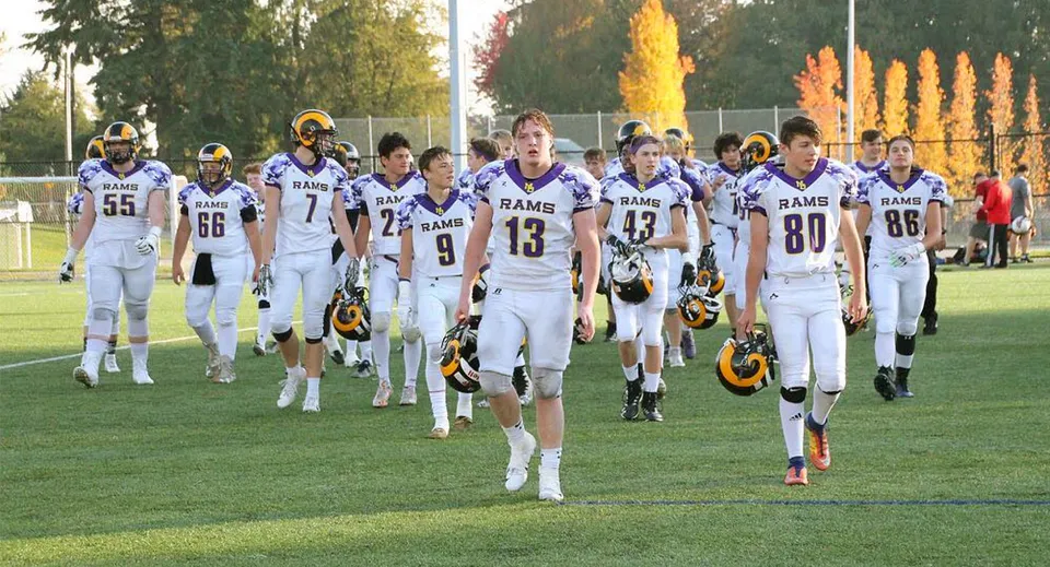 Un gruppo di giocatori di football in divisa bianca dei Rams corre sul campo della Mount Douglas Secondary School circondato da alberi autunnali.