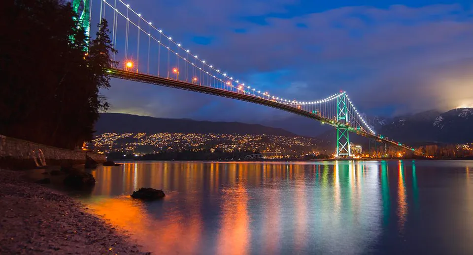 Un ponte sospeso illuminato si estende sull'acqua al tramonto con le luci della città sullo sfondo.