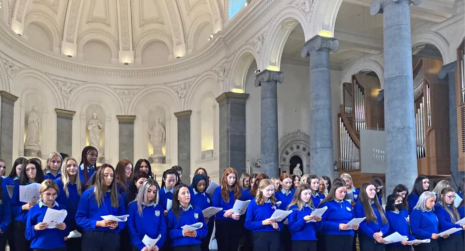 Le studentesse della Meán Scoil Mhuire in divisa blu tengono spartiti musicali in una chiesa.